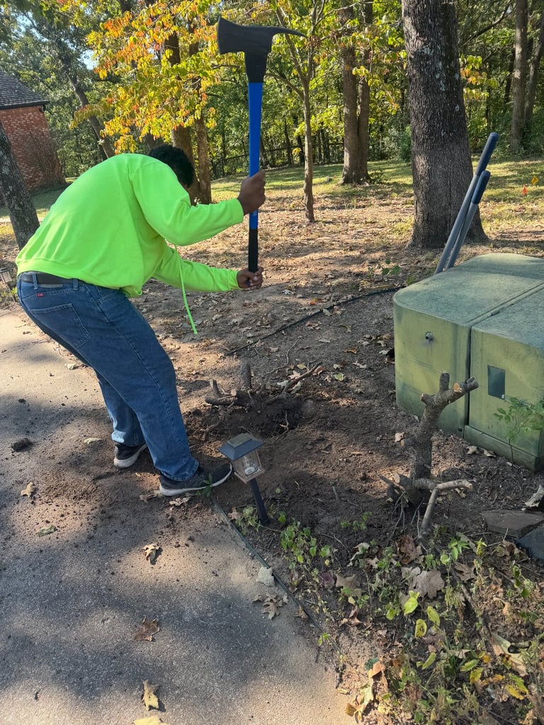 Person in green jacket using mattock to dig ground near utility box in wooded park area